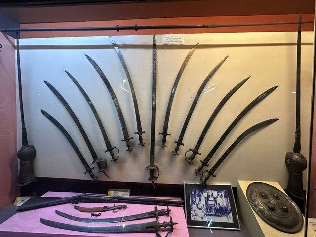 A display of curved Maratha swords fanned out across a wall, with daggers, a pistol, a shield and a period photograph below